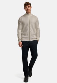 Ljusbeige zip-up tröja med fiskbensmönster, kombinerad med mörka byxor och svarta sneakers, mot en enfärgad vit bakgrund.