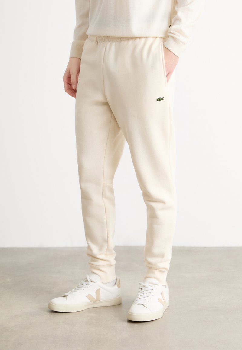 Crème-kleurige sweatpants met een elastische tailleband, zijzakken en een klein groen logo op de dij, gecombineerd met witte sneakers.
