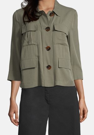 Donna che indossa una giacca verde oliva con bottoni, con quattro tasche frontali con pattina e maniche a tre quarti, abbinata a pantaloni neri.