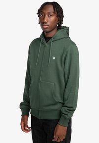 Sudadera verde con cremallera fabricada en una mezcla de algodón, que cuenta con un bolsillo frontal, capucha ajustable con cordón y un pequeño logo en el pecho.