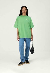 Camiseta verde de gran tamaño con mangas cortas, combinada con jeans rectos de color azul claro y zapatillas negras. Sosteniendo un pequeño bolso negro.
