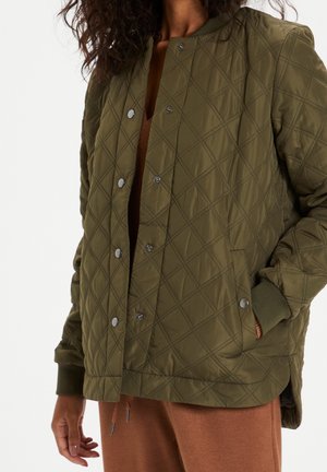 Femme portant une veste matelassée vert olive avec des boutons-pression et des poignets côtelés, sur un pantalon couleur rouille, main dans la poche.