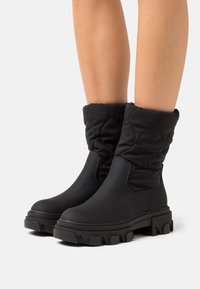 Schwarze gepolsterte Ankle Boots mit oberer Textur und dicker Gummisohle. Das Design verfügt über einen gerafften Schaft und ein grobes Profil.