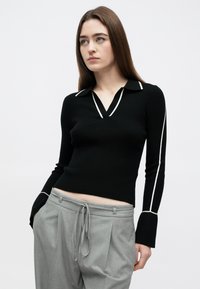 Pulôver preto em malha canelada com decote em V e acabamento branco nas golas e punhos. Usado com calças cinzentas de cintura alta.