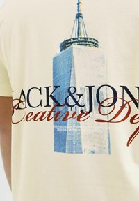 Beige puuvillane T-särk, millel on sinine graafika pilvelõhkujast, kus on suured tumesinised ja punased kirjasti "JACK & JONES" ning väiksem valge tekst.