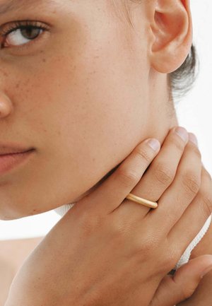 Anillo de tono dorado con una forma suave y redondeada. Se pueden ver el tono y la textura de la piel, con un fondo suavemente iluminado.