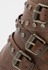 Stivale in pelle marrone con più cinturini testurizzati dotati di fibbie in tonalità argento e borchie tonde, che mettono in evidenza dettagli hardware accurati.