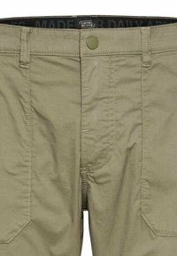 Pantalons couleur kaki avec une texture lisse, dotés d'une fermeture à bouton à l'avant, de poches latérales et de coutures contrastées. Étiquette de marque visible à la taille.