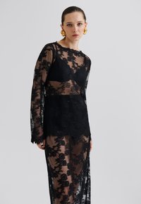 Svart blondekjole med lange ermer, prydet med blomster mønstre, en scalloped kant, og et gjennomsiktig lag. Akkompagnert av et svart undertøy.