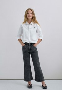 Camisa blanca de botones con las mangas remangadas, combinada con pantalones de mezclilla negros de tiro alto y campana con un patrón texturizado. Bailarinas negras completan el look.