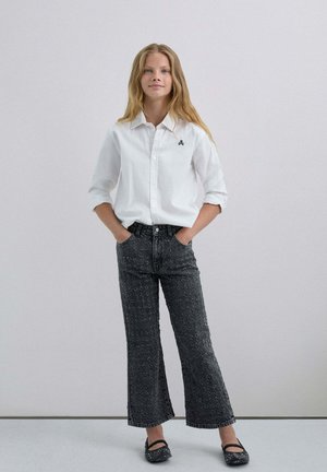 Camisa blanca de botones con las mangas remangadas, combinada con pantalones de mezclilla negros de tiro alto y campana con un patrón texturizado. Bailarinas negras completan el look.