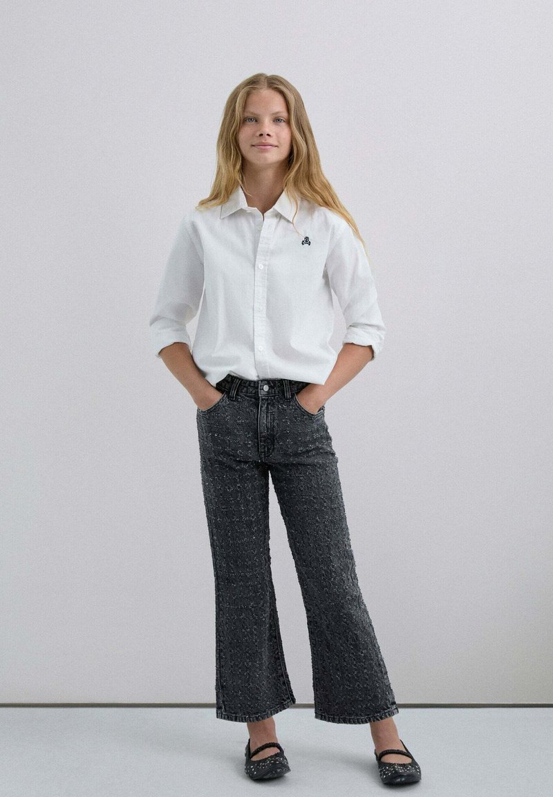 Camisa blanca de botones con las mangas remangadas, combinada con pantalones de mezclilla negros de tiro alto y campana con un patrón texturizado. Bailarinas negras completan el look.