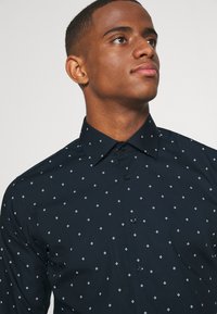 Junger Mann, der nach oben schaut, trägt ein dunkelblaues, geknöpftes Hemd mit kleinem weißem Rautenmuster auf hellem Hintergrund.