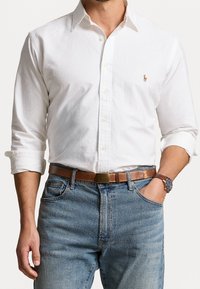 Camicia bianca con bottoni e maniche lunghe, abbinata a una cintura in pelle beige e jeans blu. Orologio al polso e la camicia presenta un logo discreto.