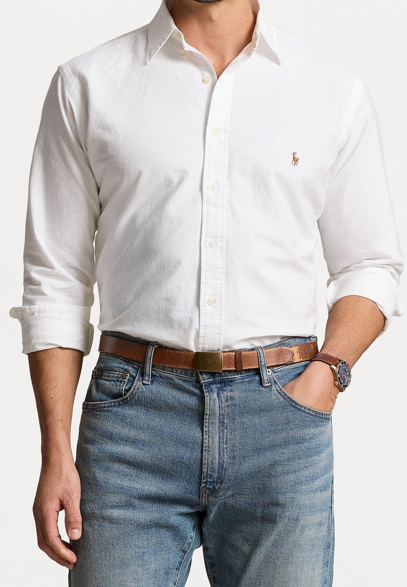 Camicia bianca con bottoni e maniche lunghe, abbinata a una cintura in pelle beige e jeans blu. Orologio al polso e la camicia presenta un logo discreto.