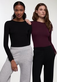 Dwie kobiety stoją obok siebie, jedna w dopasowanej czarnej bluzie z długim rękawem i szarych dresach, a druga w dopasowanej bordowej bluzie i czarnych dresach.