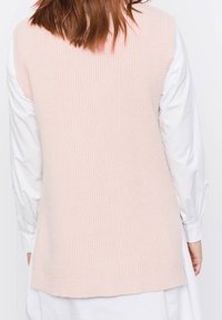 Helles rosa ärmelloses Strickoberteil mit gerippter Textur, das eine gerade Silhouette und einen abgerundeten Saum aufweist, getragen über einer weißen Langarmbluse.