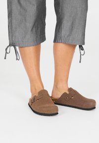 Ruske slip-on clogs lavet af tekstureret materiale med en rem og spænde detaljer. Bæres med grå cropped bukser, der er bundet ved kanten.