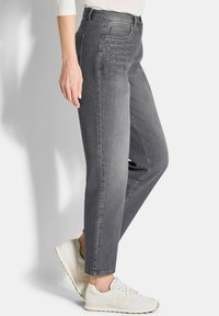 Femme portant un jean droit gris délavé avec de petites rhinestones au niveau de la poche avant et des baskets blanches.