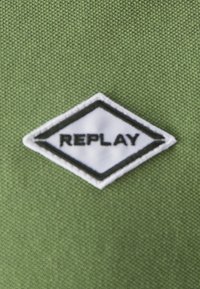 Tela verde con una superficie texturizada presenta una insignia en forma de diamante blanca, que muestra la palabra "REPLAY" en letras negras.