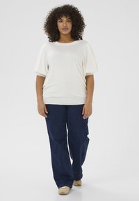 White knit top with short, puffed sleeves and a scoop neckline, paired with loose dark jeans and beige sneakers. Neutral background.