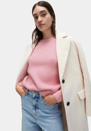 Cappotto di lana color crema con bottoni neri indossato sopra un maglione rosa testurizzato. Jeans a vita alta blu chiaro. La modella ha i capelli lunghi e castani.