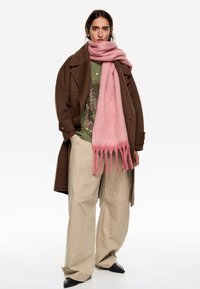 Abrigo de lana marrón, camisa verde oliva, pantalones crema de pierna ancha y bufanda de lana rosa con bordes flecados. Texturas suaves, diseño casual.