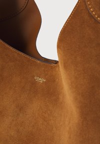 Bruine suède tas met een gladde oppervlakte, met het "COACH NEW YORK" logo in goud. De tas heeft een eenvoudig maar elegant ontwerp.