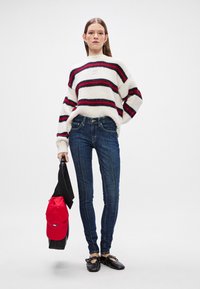 Suéter de malha às riscas em branco, vermelho e preto; jeans azuis justos; segurando uma capa vermelha; sapatos pretos; fundo minimalista.