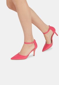 Zapatos de tacón alto de charol rosa con puntera puntiaguda y correa al tobillo. Cuentan con un tacón delgado y una textura suave y brillante.