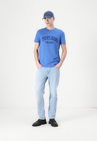 Mėlynas medvilnės marškinėliai su „TOMMY JEANS New York“ spauda, derinami su šviesiai mėlynomis džinsais ir juodais batais, papuošti mėlynu beisbolo kepure.
