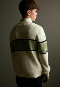 Maglione crema con texture a coste, caratterizzato da una striscia orizzontale verde e blu navy sulla schiena. Collo alto e stoffa morbida, design aderente.