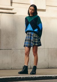 Neulottu vihreä ja sininen timanttikuviollinen neulepaita yhdistettynä laskostettuun siniseen ja keltaiseen ruutupellahameeseen. Mustat nauhalliset saappaat harmaiden sukkien kanssa.