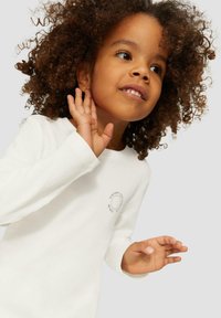 Jong kind met krullend haar, gekleed in een witte langemouwshirt, houdt hand bij het oor alsof het aandachtig luistert.