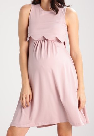 Kobieta w ciąży ubrana w różową, bezrękawnikową sukienkę z falowanym, warstwowym gorsetem oraz zdobieniem w talii.