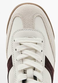 Sneaker bianca con accenti in suede, caratterizzata da un design beige e bordeaux. Parte superiore testurizzata, punta rotonda e chiusura con lacci bianchi. Suola in gomma.
