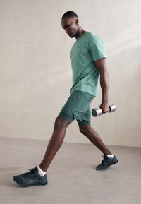 Grön atletisk t-shirt och shorts, strukturerad tyg, svarta träning skor, håller en silver hantel, i en minimalistisk inomhusmiljö.