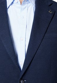 Granatowy żakiet teksturowany z jednym guzikiem, z subtelnym wzorem i jasnoniebieską koszulą na guziki pod spodem, z małym logo na klapie.