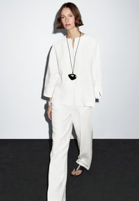 Weißes Leinenoutfit mit einem Hemd mit Knöpfen und weit geschnittenen Hosen. Ein schwarzes herzförmiges Anhänger hängt an einer schwarzen Schnur um den Hals.