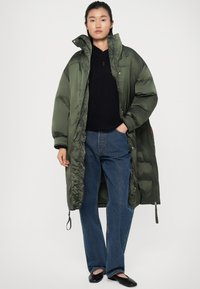 Doudoune vert olive avec un col montant et des manches surdimensionnées, dotée d'une fermeture éclair et de détails élastiques sur l'ourlet. Pull en maille noire texturée et jean bleu.