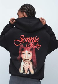Felpa nera con cappuccio che presenta una grande grafica stampata sul retro, con testo rosso che recita "Jennie Ruby" e un'immagine di una persona con lunghi capelli divisi.