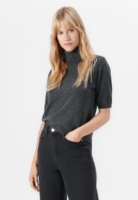 En kvinne med blondt, bølgete hår har på seg en kortermet mørk grå høyhalset genser og svarte høytlivsjeans, med hånden i lomma, mot en nøytral bakgrunn.