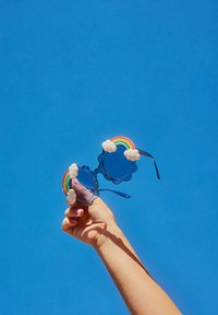 Ochelari de soare circulari, de culoare albastră, cu accente de nori și un design curcubeu pe brațe, ținuți împotriva unui cer senin de culoare albastră.