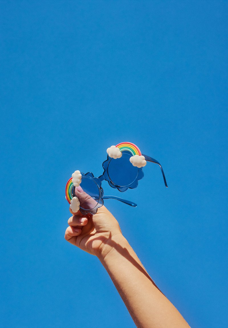 Ochelari de soare circulari, de culoare albastră, cu accente de nori și un design curcubeu pe brațe, ținuți împotriva unui cer senin de culoare albastră.