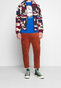 Fleecejacka i multicolor kamouflage-mönster, blå t-shirt med tryck, bruna corduroybukser, och gröna högstövlar.