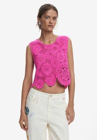 Fuchsiová háčkovaná crop top s kvetinovými vzormi a zvlneným spodným okrajom. Kombinovaná s bielymi džínsami s farebnými vyšívanými akcentmi.