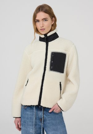 Giacca in pile color crema con colletto alto, tasca con zip nera e cerniere a contrasto nere; presenta una superficie morbida e strutturata.