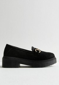 Mocasines de ante negro con un acento de metal dorado, que cuentan con una suela gruesa de plataforma negra y un detalle en zigzag a lo largo del borde.