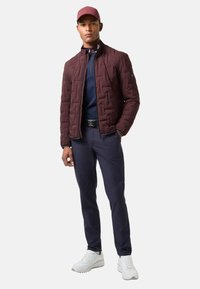 Dunkelroter gesteppter Mantel mit hohem Kragen, dunkelblaues Hemd, marineblaue Hose und weiße Sneaker, mit tailliertem Schnitt und dezenten Hardware-Akzenten.