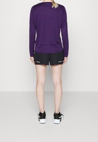 Persona de pie de espaldas, vistiendo una camiseta de manga larga morada, pantalones cortos deportivos negros y zapatillas negras y blancas sobre un fondo liso.